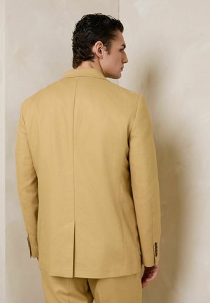 Oldalról néző férfi, aki bézs öltönyzakót és hozzáillő nadrágot visel, egy sima bézs fal előtt.