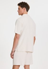 Chemise beige clair à manches courtes et shorts assortis en tissu texturé. La chemise possède un col et des fentes latérales pour plus de confort.