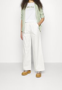 Pantaloni bianchi a gamba larga, camicia verde con bottoni sopra una maglietta bianca con la scritta "BOSS" in lettere verdi, e scarpe beige con suola spessa.