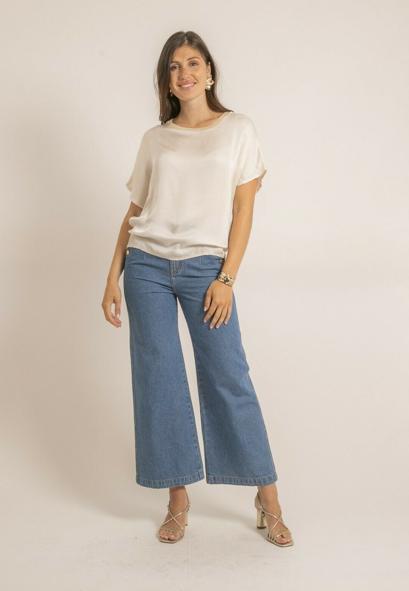 Haut léger beige clair, transparent, à coupe ample, associé à un jean en denim bleu à jambes larges. Le modèle porte des talons à lanières et un bracelet. Design simple et minimal.