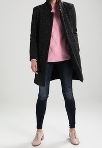 Cappotto lungo nero con trama, sopra un maglione rosa a trecce, abbinato a jeans skinny scuri e scarpe stringate rosa chiaro.