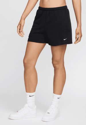 Persoon draagt zwarte Nike-short, witte Nike-sokken en witte sneakers, staat met ontspannen armen langs het lichaam tegen een effen achtergrond.