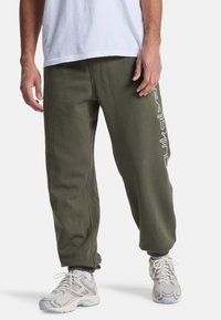 Olivgröna sweats med avslappnad passform, elastiska mudd och en vit logodetalj på sidan. Matchas med ljusa sneakers.