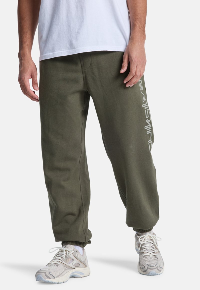 Olivgröna sweats med avslappnad passform, elastiska mudd och en vit logodetalj på sidan. Matchas med ljusa sneakers.