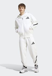 Survêtement de sport blanc avec une veste à capuche zippée et un pantalon assorti. Comprend des accents noirs et le logo Adidas sur la poitrine et la cuisse.