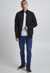 Veste zippée noire sur une chemise blanche, associée à un jean bleu. La veste a un col haut et des poignets côtelés. Des baskets noires complètent la tenue.