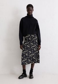 Gestrickter schwarzer Rollkragenpullover, schwarzer Rock mit Blumenmuster und weißen Akzenten, kombiniert mit schwarzen Schuhen und Socken, vor einem neutralen Hintergrund stehend.