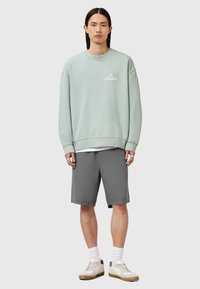 Sudadera de cuello redondo verde menta con logo blanco, combinada con pantalones cortos grises. El modelo lleva zapatillas blancas con suelas color beige claro y calcetines blancos.