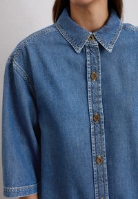 Hellblaue Jeansbluse mit Knopfleiste, Kragen und kurzen Ärmeln. Mit goldenen Knopfgriffen und sichtbaren Nähten.
