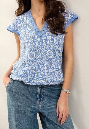 Vrouw die een blauw-witte blouse met bloemenpatroon en V-hals draagt, afgewerkt met haakwerk, gecombineerd met een blauw, hooggetailleerd spijkerbroek en een zilveren armband.