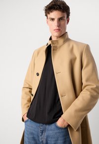 Palton bej din lână cu guler înalt, închidere cu butoane în față și detalii discrete de cusătură, purtat peste un tricou negru și blugi albaștri.