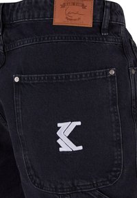 Sorte denim-bukser har et lærredsskilt i taljen, en baglomme med hvid broderet logo og sølv hardware-detaljer.