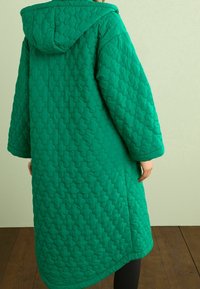 Manteau long matelassé vert avec capuche, présentant des motifs hexagonaux et une coupe décontractée. Texture lisse et manches longues complètent le design.