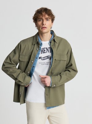 Mężczyzna poprawiający zieloną kurtkę na guziki na niebieską koszulę dżinsową i białą koszulkę z nadrukiem, stojący na tle jednolitego jasnego tła.