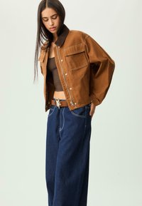 Kvinde iført en brun cropped jakke, mørkebrun tank top, bredbenede mørkeblå jeans og et bælte med en stjernet spænde, mens hun kigger ned.