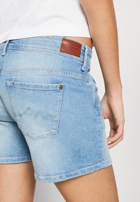 Ljusblå denimshorts med en sliten yta, med en brun läderbrandpatch, fem fickor och en uppvikt fåll.