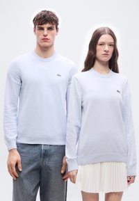 Νέος άνδρας και γυναίκα στέκονται δίπλα δίπλα φορώντας ανοιχτό μπλε πουλόβερ με πράσινα λογότυπα κροκοδείλου, ο άνδρας με τζιν, η γυναίκα με άσπρη πλισέ φούστα.