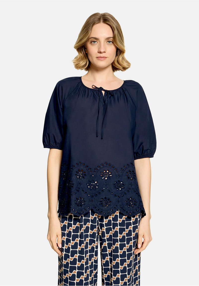 Femme aux cheveux blonds courts portant un chemisier bleu marine avec des détails en œillets et un pantalon large à motifs sur un fond clair uni.