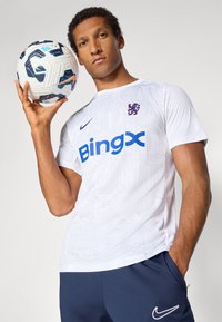 Hvit kortermet skjorte med preget mønster, fremtredende blå "Bingx"-logo, marinblå shorts og en mønstret fotball holdt i hånden.