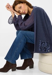 Shawl lavorato a maglia blu navy con una tasca accentuata da paillettes, abbinato a jeans a zampa d'elefante in denim scuro e stivaletti alla caviglia in suede marrone.