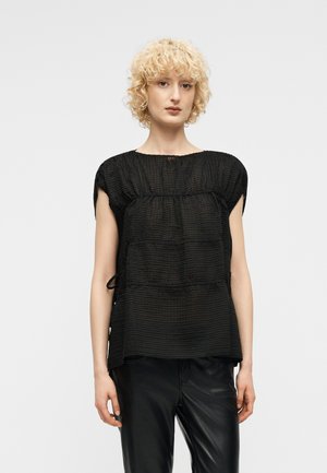 Ξανθιά γυναίκα με κοντά, σγουρά μαλλιά, που φοράει μαύρο μπλουζάκι με υφή και κοντά μανίκια και μαύρο δερμάτινο παντελόνι, στέκεται μπροστά σε απλό φόντο.