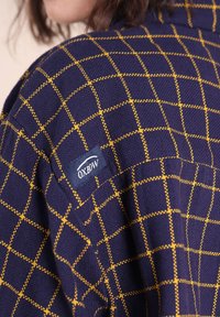 Personne portant un pull violet à motif de carreaux jaunes avec une étiquette de la marque Oxbow sur la manche supérieure.