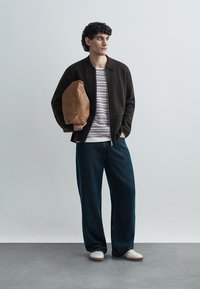 Dunkelbraune Jacke mit Reißverschluss, gestreifter weiß-blauer Pullover, locker sitzende dunkle Jeans, beige Sneaker, hält eine braune Tasche mit Struktur.