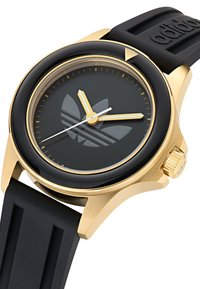 Zeltītas krāsa pulkstenis ar melnu ciparnīcu, kurā ir trīs rādītāji, zeltīti akcenti un melna gumijas siksna. Ciparnīcā ir redzama ievērojama logo motīvs.