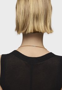 Collar metálico delgado en un color claro, con un diseño minimalista y un pequeño acento central, que se lleva con una blusa negra sin mangas.