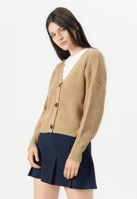 Strikket cardigan i lysebrun med V-hals, knapper nedover foran og ribbestrikket tekstur. Bæres over en hvit topp og marineblå skjørt.