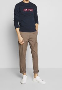 Námořnická mikina s červeným logem "KORS", béžové chino kalhoty s ohrnutými nohavicemi a bílé tenisky. Směs bavlny, volný střih a minimalistický design.