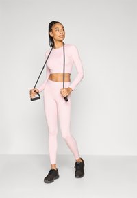 Ροζ αθλητικό σετ που περιλαμβάνει μακρυμάνικο cropped τοπ και εφαρμοστά κολάν, και τα δύο από υλικό με λεία υφή. Συμπεριλαμβάνονται μαύρα αθλητικά παπούτσια και λάστιχο αντίστασης.