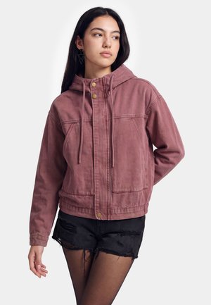 Jonge vrouw met lang donker haar die een mauve capuchonjas, zwarte gescheurde shorts en doorzichtige zwarte panty's draagt, kijkt opzij.