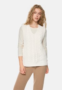 Young woman with wavy blonde hair wearing a white cable-knit sleeveless sweater over a cream long-sleeve shirt and beige knit pants.