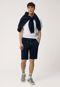 Marineblauwe shorts en een wit poloshirt, met een marineblauwe sweatshirt over de schouders gedrapeerd. Met een trekkoord in de taille, gestreepte sokken en dikke sneakers.