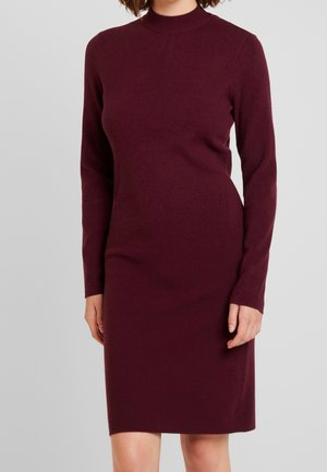 Femme portant une robe en maille bordeaux ajustée, à manches longues, arrivant au genou, avec un col haut, debout devant un fond gris uni.