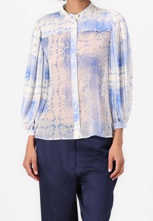 Kobieta ubrana w przezroczystą, zapinaną na guziki bluzkę z niebieskim kwiatowym wzorem i plisowanymi, balonowymi rękawami, w połączeniu z ciemnoniebieskimi, wysokim stanem spodniami.