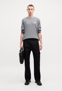 Γκρι μπλουζάκι με μακριά μανίκια και μικρό λογότυπο, μαύρο παντελόνι με πλαϊνές τσέπες και μαύρα παπούτσια. Το μοντέλο κρατά μια μαύρη τσάντα.