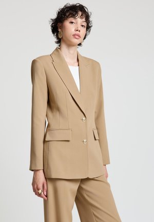 Femme aux cheveux courts et bouclés portant de grandes boucles d'oreilles en or, un blazer beige cintré avec des boutons dorés et un pantalon beige assorti sur un fond uni.