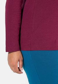 Maroon long-sleeve top with a straight hem, paired with teal leggings. The fabric appears soft, showing a smooth texture and clean stitching.