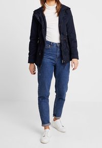 Marineblaue Kapuzenjacke mit Reißverschluss und Knopfverschluss, kombiniert mit hellblauen Jeans. Das Modell trägt weiße Sneakers.