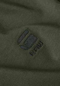 Maslinasto zelena tkanina s glatkom teksturom, koja sadrži crni izvezeni logo i tekst na površini. Jednostavan dizajn i jednobojna boja.