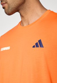 Φωτεινή πορτοκαλί βαμβακερή μπλούζα με στρογγυλή λαιμόκοψη, κοντά μανίκια και ένα ναυτικό λογότυπο με τη λέξη «TENNIS» στην αριστερή πλευρά του στήθους.