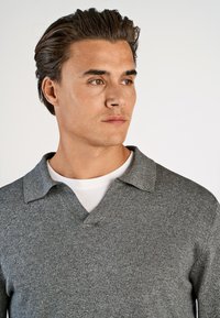 Mand med kort mørkt hår iført en grå strikket sweater med krave over en hvid t-shirt, som ser til venstre på en ensfarvet baggrund.