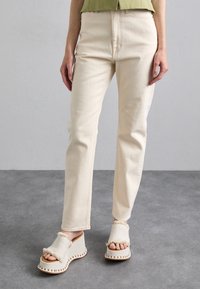 Beige bomullbyxor med rak passform och subtil struktur, kombinerat med ljusa kilsandaler som har fransiga detaljer och kontrasterande sömmar.