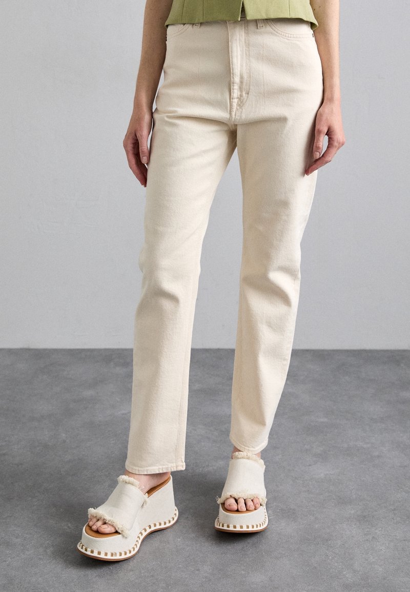 Beige bomullbyxor med rak passform och subtil struktur, kombinerat med ljusa kilsandaler som har fransiga detaljer och kontrasterande sömmar.