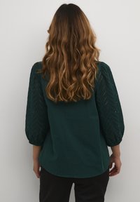 Temno zelena majica s tremi četrtnjami dolgimi rokavi, ki vsebuje kvačkanje v črno-beli črti. Tkanina se zdi mehka in lahka.