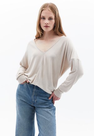 Beige Strickpullover mit V-Ausschnitt, lockerer Passform und langen Ärmeln. Kombiniert mit hochgeschnittenen blauen Jeans. Weicher Stoff, lässiger Stil.