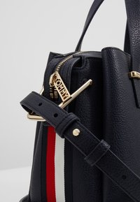 Sac à main en cuir noir texturé avec une bande rouge, blanche et noire, une fermeture éclair dorée et une bandoulière noire réglable avec boucle.