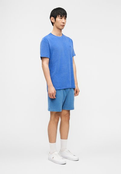 Άνδρας όρθιος, φοράει γαλάζιο κοντομάν ικο πουκάμισο και αντίστοιχα σορτς με λευκές κάλτσες και λευκά αθλητικά παπούτσια, μπροστά σε λευκό απλό φόντο.